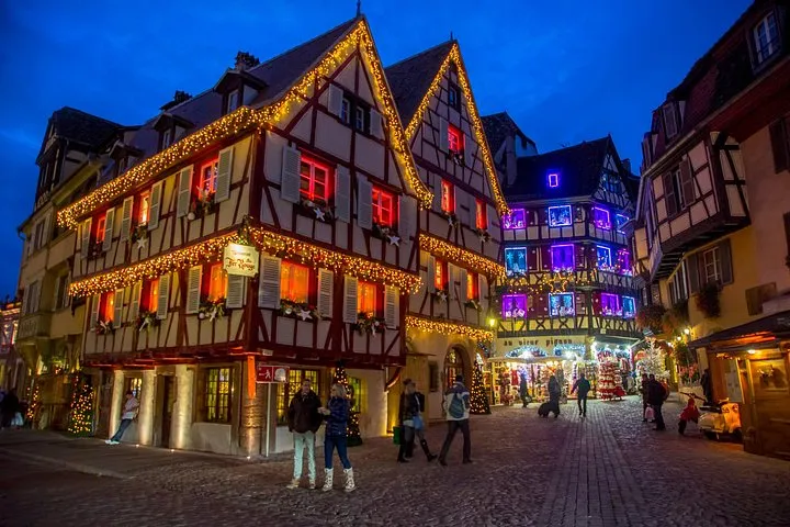A row of half-timbered gables outlined in gold lights at blue hour on a cobbled square in Colmar