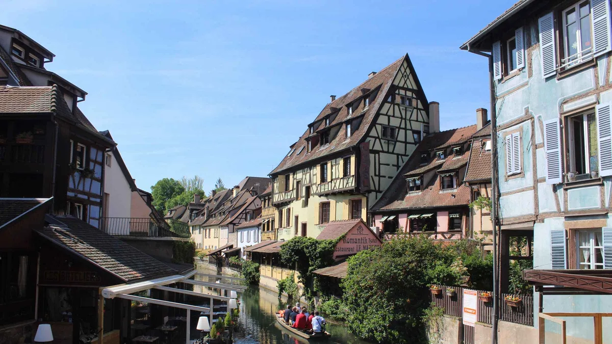 Pastel half-timbered houses along a canal in Colmar under a clear blue sky, a small boat on the water
