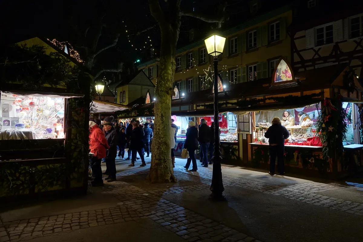 Visitors at lit market stalls under a street lamp on Place des Dominicains in Colmar